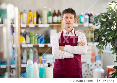 Man in apron stands with arms crossed over chest among shelves and storefronts in household store 132764224