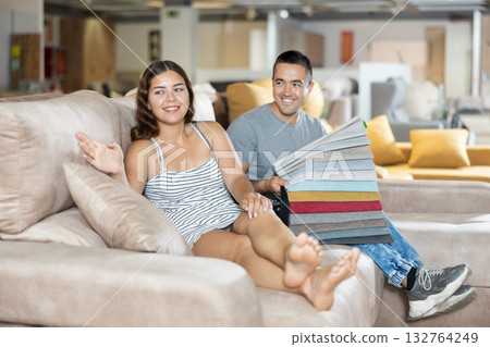 In store wife sit with legs on gray sofa to test in use,husband examines upholstery fabric 132764249
