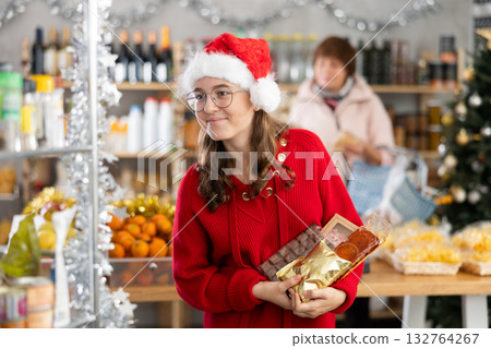 Positive girl in festive hat choosing sweets for New Year's holiday in supermarket with mother choosing food on shelves in 132764267