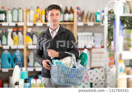 Positive man selects various household cleaning products for cleaning the house with full basket of various purchases Positive man selects various household cleaning products for cleaning the house with full basket of various purchases 132764281