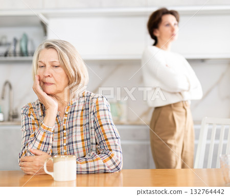 Upset old woman sitting at the kitchen table with her back to woman quarreling to her 132764442