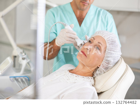 Cosmetologist performing anti-aging face hardware therapy procedure for aged female client in clinic of aesthetic medicine 132764482