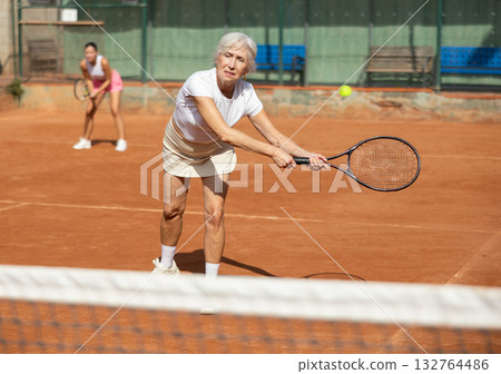 Sporty two women during doubles match on tennis court. Active old lady hitting tennis ball with racket Sporty two women during doubles match on tennis court. Active old lady hitting tennis ball with racket 132764486