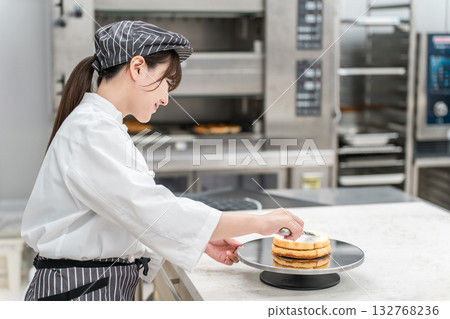 一位身穿廚師服的年輕亞裔女性（糕點師）在糕點工作坊、蛋糕店廚房或麵包房工作。 132768236