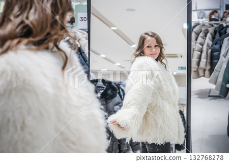 Young woman tries on a stylish white sheepskin coat in a clothing boutique while checking her reflection in the mirror during a seasonal shopping trip Young woman tries on a stylish white sheepskin coat in a clothing boutique while checking her reflection in the mirror during a seasonal shopping trip 132768278