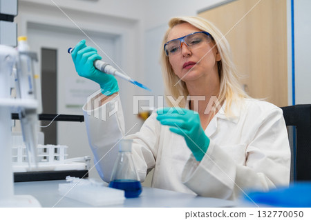 Middle aged blonde chemist in laboratory carefully preparing solution in test tube with pipette. 132770050