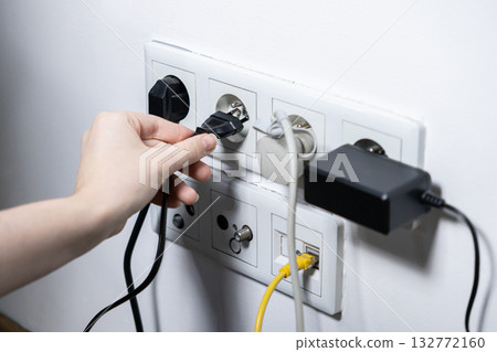 Person's hand carefully plugging a black electrical cord into a white multi-socket wall outlet, illustrating modern home power supply and connectivity 132772160