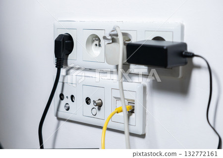 Busy wall outlet with multiple electrical plugs, network cables, and various charging adapters connected, showcasing essential modern home technology connectivity and digital energy needs Busy wall outlet with multiple electrical plugs, network cables, and various charging adapters connected, showcasing essential modern home technology connectivity and digital energy needs 132772161