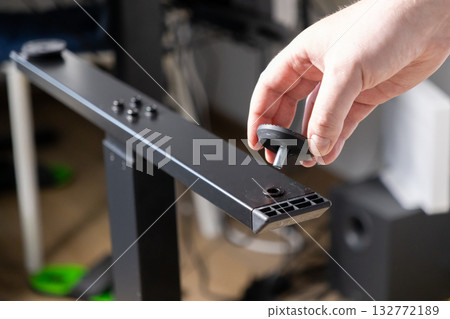 Hand holds a black adjustable leveling foot with a screw, preparing its installation onto a metal furniture leg for DIY assembly, ensuring stability 132772189