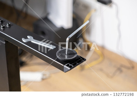 Allen key is meticulously adjusting a modern desk leg leveler, accompanied by a small wrench, signifying precise furniture assembly and practical home improvement 132772198