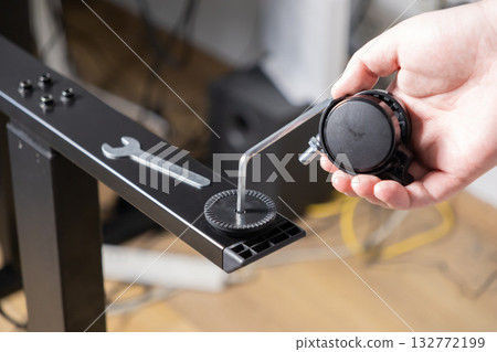 Hand assembling adjustable black metal desk frame with an Allen wrench, installing caster wheel, promoting furniture setup and DIY home office Hand assembling adjustable black metal desk frame with an Allen wrench, installing caster wheel, promoting furniture setup and DIY home office 132772199