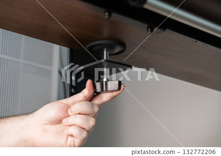 Person's hand tightening a sturdy desk clamp, installing an accessory underneath a wooden table for an organized home office workspace setup 132772206