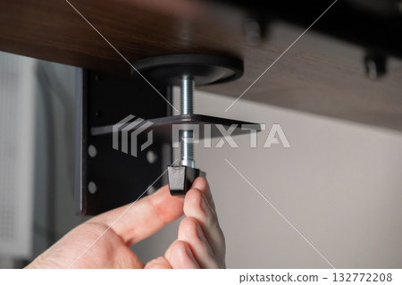 Hand adjusting black clamp with threaded screw, securely mounting device to wooden desk, demonstrating precise DIY installation for adaptable ergonomic office setup Hand adjusting black clamp with threaded screw, securely mounting device to wooden desk, demonstrating precise DIY installation for adaptable ergonomic office setup 132772208