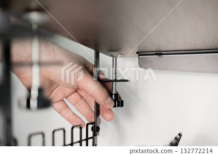 Human hand precisely adjusts metal clamp securing modern wire organizer basket under wooden desk, improving workspace efficiency and cable management Human hand precisely adjusts metal clamp securing modern wire organizer basket under wooden desk, improving workspace efficiency and cable management 132772214