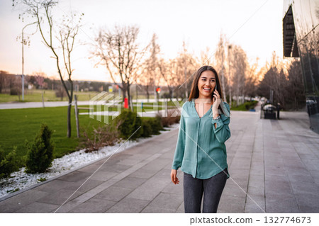 Young businesswoman walking and talking on smartphone in a park near office buildings Young businesswoman walking and talking on smartphone in a park near office buildings 132774673
