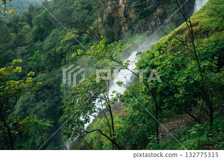 A beautiful waterfall cascades down from a rocky cliff amidst lush greenery and mist at the base of the waterfall. A beautiful waterfall cascades down from a rocky cliff amidst lush greenery and mist at the base of the waterfall. 132775315