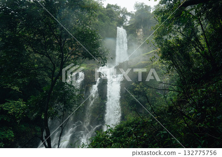 A beautiful waterfall cascades down from a rocky cliff amidst lush greenery and mist at the base of the waterfall. A beautiful waterfall cascades down from a rocky cliff amidst lush greenery and mist at the base of the waterfall. 132775756