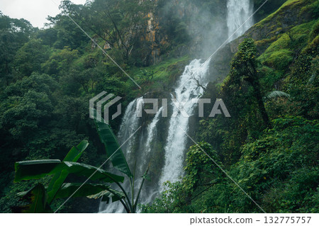 A beautiful waterfall cascades down from a rocky cliff amidst lush greenery and mist at the base of the waterfall. A beautiful waterfall cascades down from a rocky cliff amidst lush greenery and mist at the base of the waterfall. 132775757