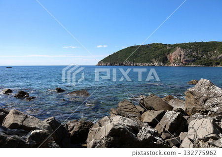 Clear blue waters and rocky coastline under a bright sky at midday near a lush green hillside Clear blue waters and rocky coastline under a bright sky at midday near a lush green hillside 132775962