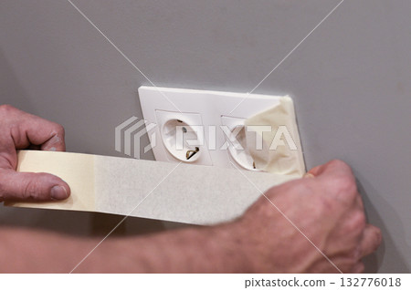 A man's hands are shown in close-up as he carefully applies adhesive tape to cover an electrical outlet, protecting it from paint splatters during a renovation project. 132776018