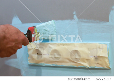 Applying paint to the wall surrounding an electrical socket using a brush, with masking tape for protection, as part of a DIY home improvement project, ensuring clean edges and safe interior upkeep. 132776022
