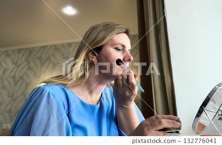 Standing before her bedroom mirror, a young woman carefully applies different makeup products to achieve her desired look. 132776041