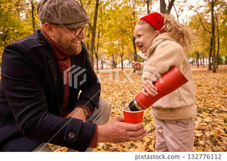 Father watching daughter pour tea into cup in autumn park Father watching daughter pour tea into cup in autumn park 132776112