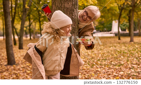 Grandmother offering sandwich to granddaughter behind tree in autumn park 132776150