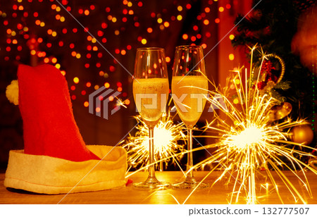 Two champagne flutes with Santa hat and sparklers on wooden table. Night festive holiday celebration with twinkling lights and decorated Christmas tree with bokeh lights 132777507