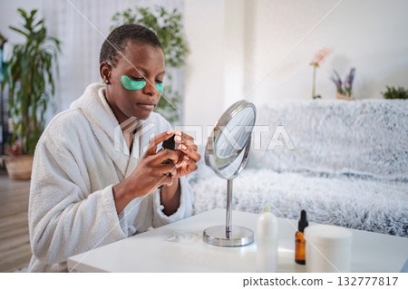 Young woman applying moisturizing serum on her face while wearing a bathrobe and under eye patches Young woman applying moisturizing serum on her face while wearing a bathrobe and under eye patches 132777817