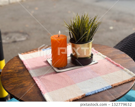 Orange candle and potted plant on a cafe terrace table. Simplicity, light and aesthetic calm in outdoor lifestyle design. Orange candle and potted plant on a cafe terrace table. Simplicity, light and aesthetic calm in outdoor lifestyle design. 132779632