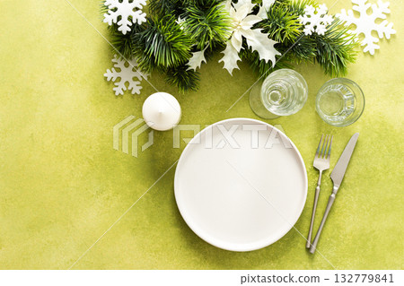 A top view of a Christmas table setting featuring an empty plate with candle and cutlery, festive spruse and white tree decorations, on a bright green background A top view of a Christmas table setting featuring an empty plate with candle and cutlery, festive spruse and white tree decorations, on a bright green background 132779841