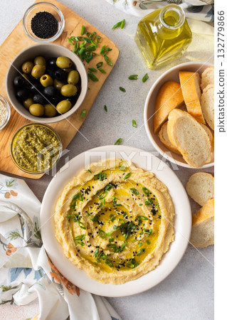 Creamy hummus is beautifully plated with green herbs and olive oil, surrounded by fresh olives, pita bread, and various seasonings on a simple table 132779866