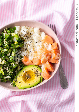 Fresh breakfast bowl featuring salmon, sliced avocado, leafy greens, and rice served on a pink striped tablecloth. Perfect for a nutritious start to the day 132779897