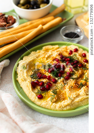 Delight in a classic hummus dish topped with pomegranate seeds, served alongside a variety of cheeses, nuts, and breadsticks perfect for sharing during gatherings 132779906