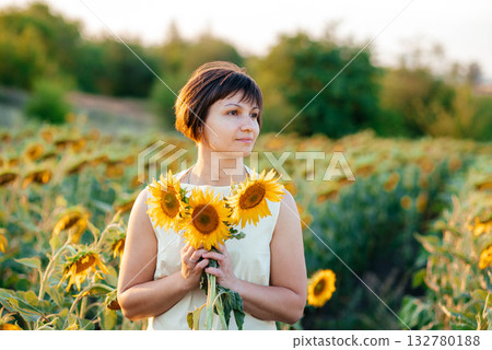 Beautiful brunette in a field of sunflowers. Girl in white in a field of sunflowers enjoys nature and herself.  132780188