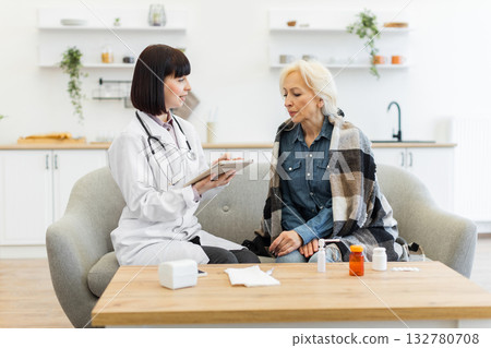 A female doctor in a white coat consults with an elderly patient wrapped in a blanket, discussing health concerns while sitting on a couch. 132780708