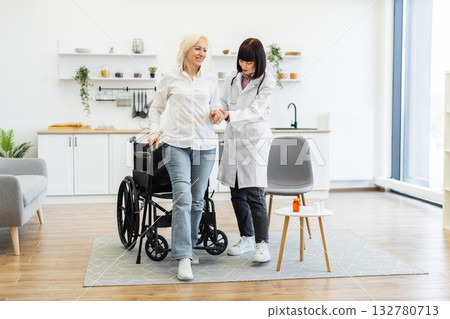 A female doctor helps an elderly woman stand up from her wheelchair in a bright room, providing support and care. A female doctor helps an elderly woman stand up from her wheelchair in a bright room, providing support and care. 132780713