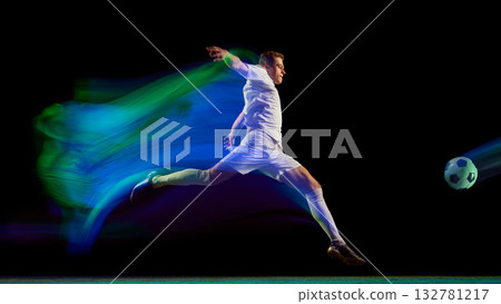 Soccer player kicking ball with green and blue neon motion trail on dark field 132781217