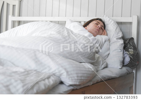 A woman is sleeping peacefully in her bed with a white pillow and a blanket. A beautiful young happy lady is sleeping peacefully on a cozy bed in the morning light at home, in a relaxed atmosphere A woman is sleeping peacefully in her bed with a white pillow and a blanket. A beautiful young happy lady is sleeping peacefully on a cozy bed in the morning light at home, in a relaxed atmosphere 132783393