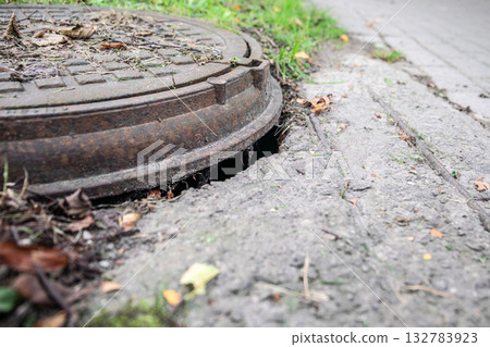 A rusty manhole cover slightly lifted from the concrete pavement, leaving a dangerous gap at the A rusty manhole cover slightly lifted from the concrete pavement, leaving a dangerous gap at the 132783923