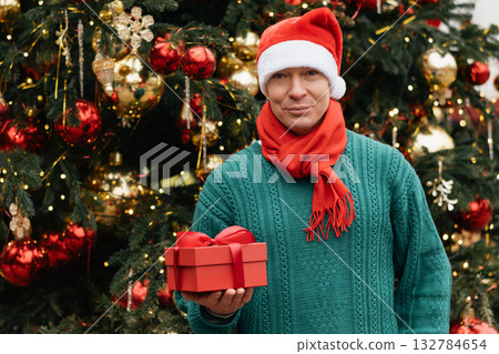 Winter portrait of happy man wearing green sweater holding and Santa Hat red gift and stand near Christmas tree. Xmas Holiday. Close up. 132784654