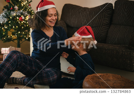 Mother And Child Share Christmas Moment, Decorating Hats By The Tree In A Cozy Living Room Mother And Child Share Christmas Moment, Decorating Hats By The Tree In A Cozy Living Room 132784863