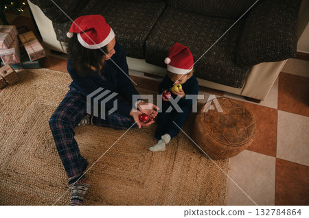 Family Christmas Moment: Mother And Child In Santa Hats Sharing Ornaments On Cozy Rug Family Christmas Moment: Mother And Child In Santa Hats Sharing Ornaments On Cozy Rug 132784864