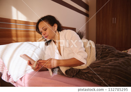 Woman Relaxing in Bed Using Smartphone, White Robe, and Cozy Blanket by Wooden Headboard 132785295