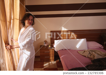 Woman In White Robe Opening Curtains In Sunlit Bedroom - Cozy Morning At Home 132785306