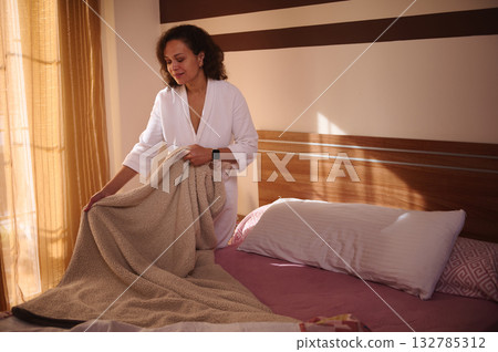 Woman In White Bathrobe Making Bed In Sunny Bedroom With Cozy Blanket 132785312