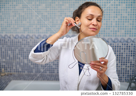 Woman In White Robe Applies Earring In Front Of Mirror During Personal Self-Care 132785330