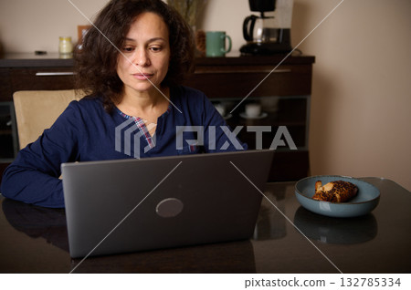 Woman Working On Laptop At Dining Table With Croissant On Plate Woman Working On Laptop At Dining Table With Croissant On Plate 132785334
