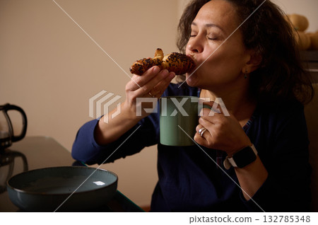 Woman Enjoys Warm Pastry While Sipping Coffee From A Green Mug In Kitchen Woman Enjoys Warm Pastry While Sipping Coffee From A Green Mug In Kitchen 132785348
